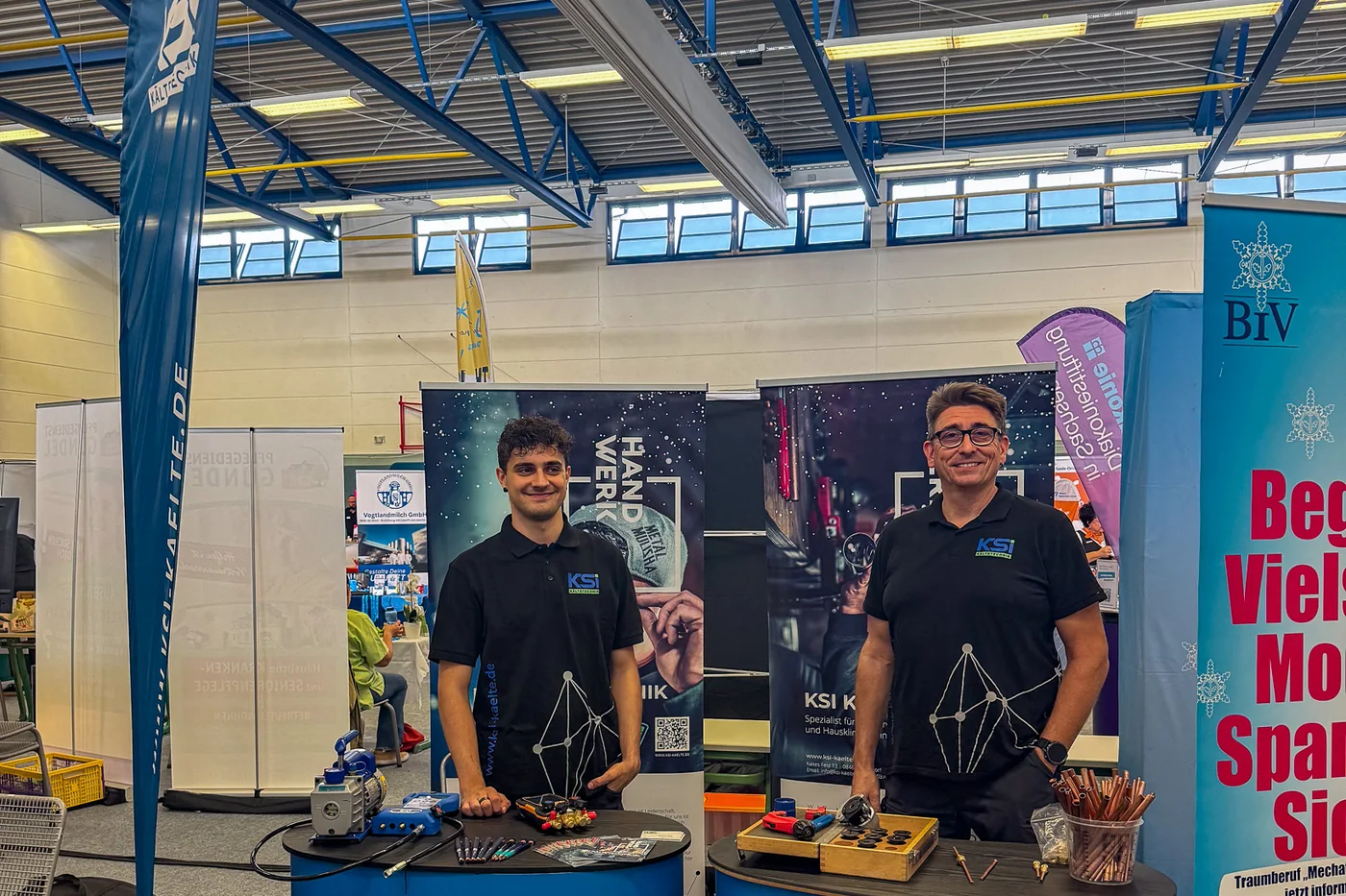 The KSI Kältetechnik exhibition stand in the sports hall on Cunsdorfer Straße in Reichenbach, with two staff members behind the blue counters